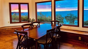 Uma área de refeições com mesa e cadeiras de madeira, janelas amplas com vista para o mar e um lustre.
