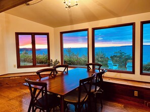 A dining area with a wooden table and chairs, large windows with a view of the sea, and a chandelier.