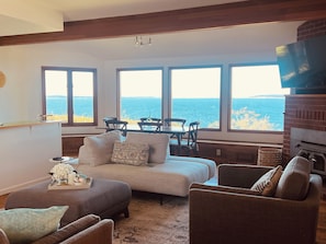 A cozy living room with a sofa, armchairs, a fireplace, and a view of the sea through large windows.