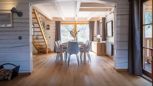 A spacious dining area with a wooden table and chairs, a staircase, and large windows offering a view of the outdoors. - Chalet Choquette - secluded family break for 7 with spa - OVO Network (Les Houches)