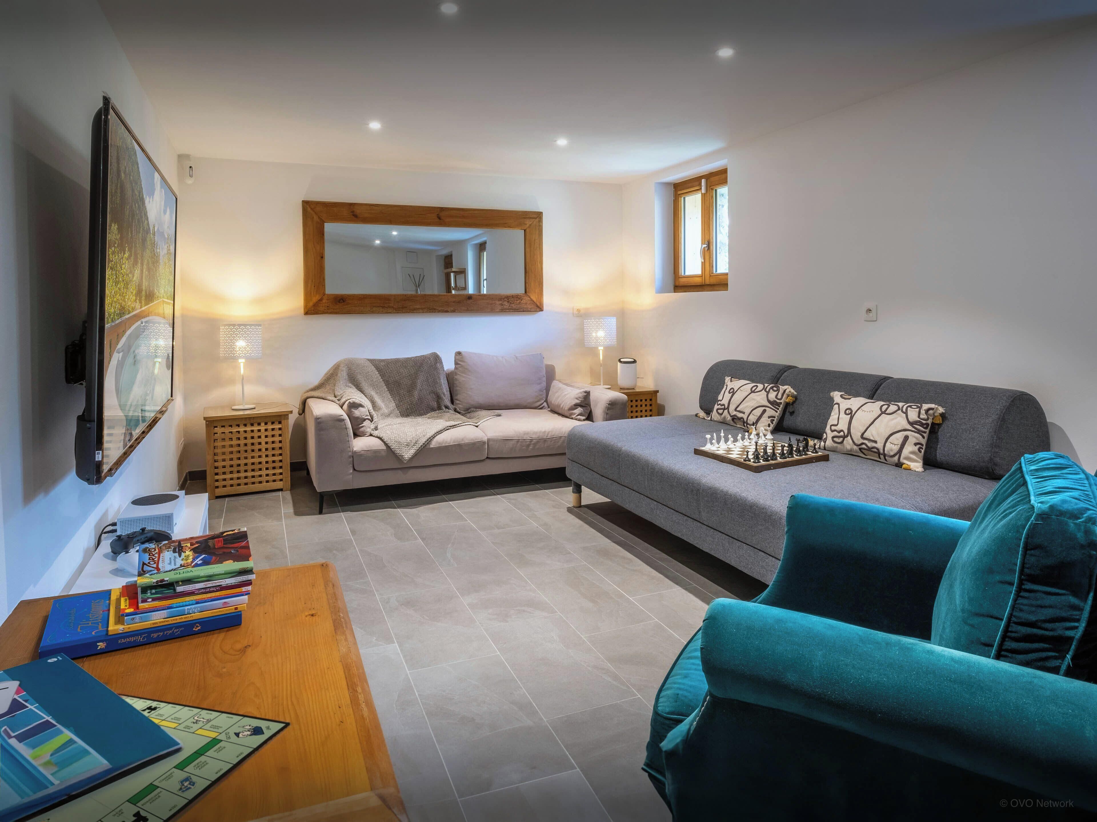 A living room with two sofas, a coffee table with books, a chessboard, and a TV mounted on the wall.