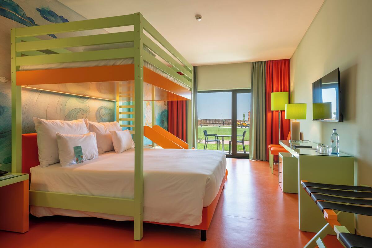 A hotel room with a bunk bed, a desk with a chair, a TV, and a window with a view of the outdoors.