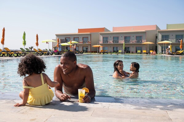 Zona de piscina con sillones de descanso y sombrillas, un edificio con balcones y gente relajándose junto a la piscina.