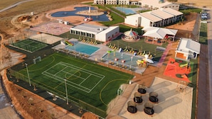 Vue aérienne d’un complexe hôtelier comprenant plusieurs bâtiments, une grande piscine, un court de tennis et une aire de jeux.