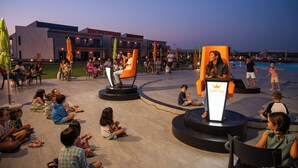 Un espace extérieur aménagé avec des sièges, où des personnes sont rassemblées autour d’un podium. On aperçoit en arrière-plan un bâtiment moderne et, sur la droite, une piscine.