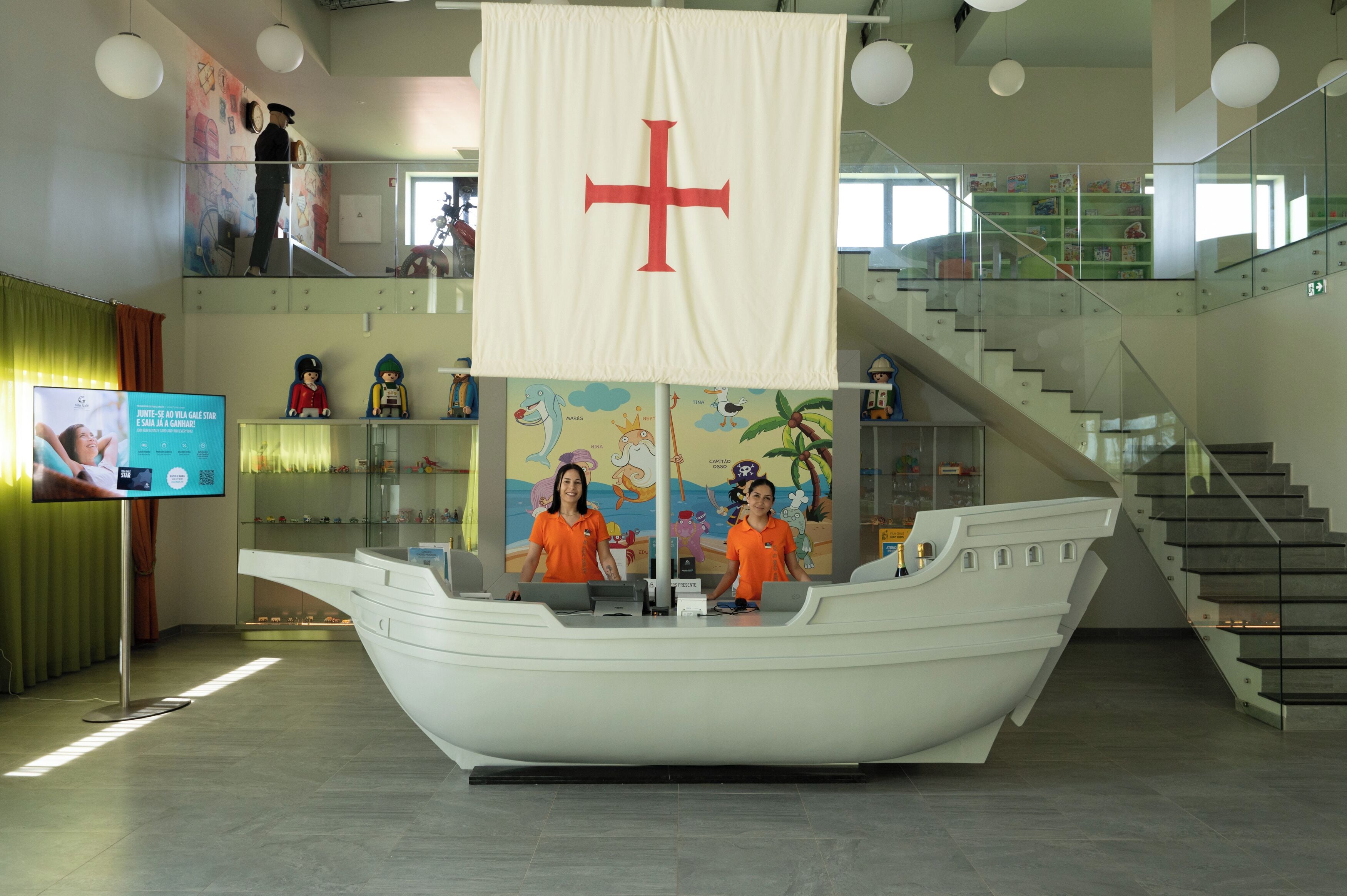 Un área de recepción con una barra con forma de barco, un gran cartel con una cruz roja y una escalera con barandillas de vidrio.