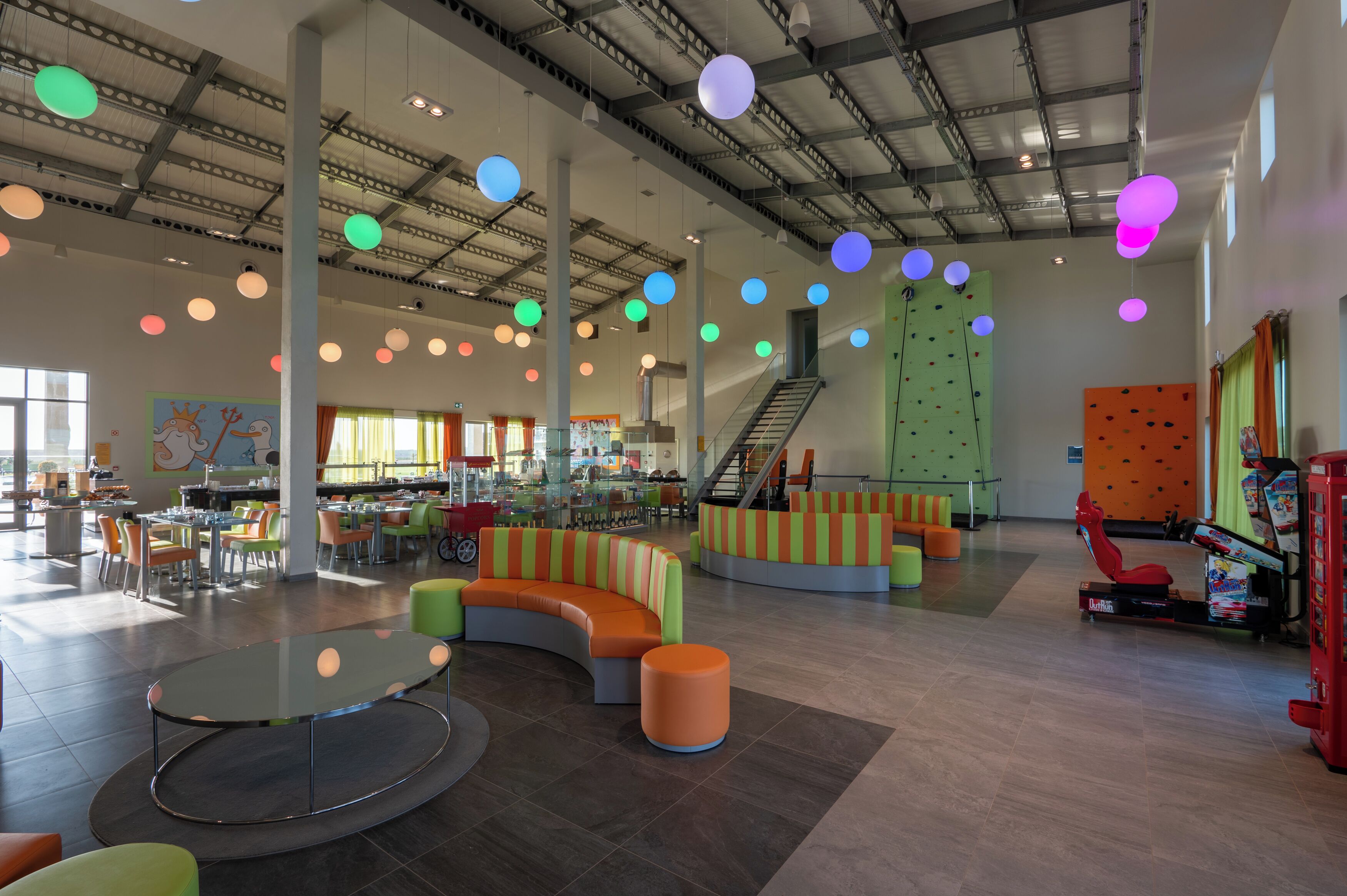 A spacious indoor area with high ceilings, colorful hanging spherical lights, a central seating area with a curved sofa and ottomans, a staircase, and a climbing wall.