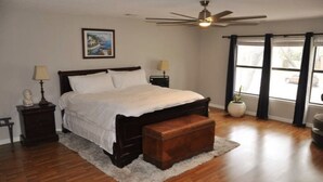 Un dormitorio con cama, ventilador de techo, ventana con cortinas, mesita de noche, lámpara y un cuadro en la pared.