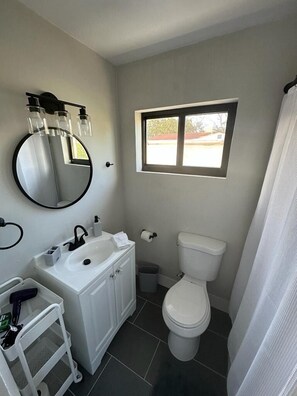 A compact bathroom with a white sink, mirror, and toilet. A window provides natural light, and a small trash bin is visible. - Charming Miami Studio (Miami)