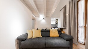 A modern living room with a grey sofa, wooden beams on the ceiling, and a kitchen in the background. - Modern Gem in Castello (Cagliari)