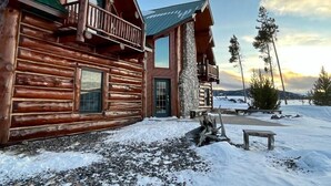 A log cabin with a balcony and a stone chimney, surrounded by snow and trees. - The Wyoming Lodge and Game Room (Dubois)