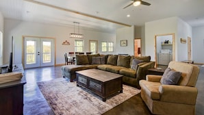 A spacious living room with a large sectional sofa, a wooden coffee table, and a ceiling fan. There is a dining area with a table and chairs, and a kitchen area with a refrigerator and cabinets. - Lakefront Retreat w/Views, Fire Pit & 2 Acres (Golden)