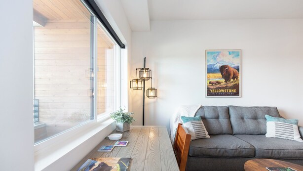 A modern living room with a grey sofa, a wooden coffee table, and a large window. There is a poster on the wall and a floor lamp.