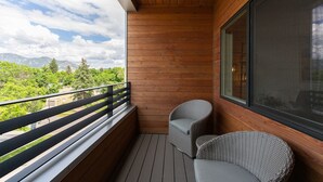 A balcony with wooden flooring, two wicker chairs, and a large window offering a view of greenery and mountains.