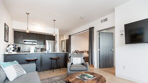 A modern living room with a grey sofa, a round coffee table, and a kitchen area with dark cabinets and a built-in oven.