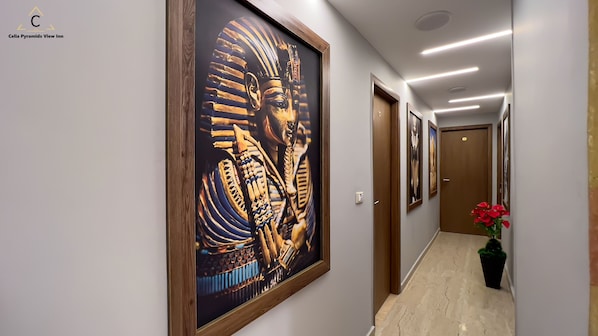 A hallway with a large framed artwork of an Egyptian figure, a wooden door, and a potted plant. - Celia Pyramids View inn (Giza)
