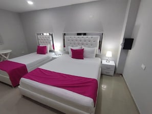 Hotelkamer met twee bedden, elk met een witte hoofdbord en roze beddengoed. Er staan nachtkastjes met lampen, een klein wit tafeltje en er is een tv aan de muur bevestigd.