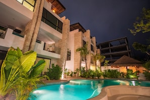 A modern hotel with a pool, surrounded by tropical plants and a thatched-roof structure.