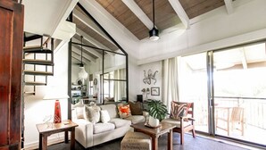 A spacious living room with a large sofa, coffee table, and a dining area. There is a staircase leading to an upper level, and a balcony with outdoor seating.