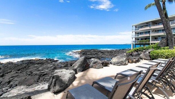 A beachside view with a rocky shoreline, lounge chairs, and a building with balconies overlooking the ocean.