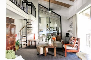 Relax in a spacious living area with a high ceiling and exposed beams, featuring a large open-plan space with a sofa, armchairs, and a staircase leading to an upper level.