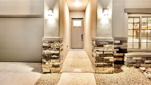 A modern house entrance with a stone facade, a concrete pathway, and a garage door on the left. - Modern Accessorized 3-Bedroom (Las Cruces)