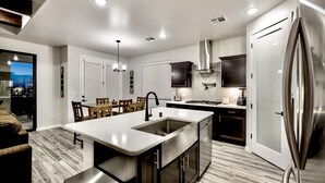 A modern kitchen with a central island, stainless steel appliances, and a dining area with wooden chairs. - Modern Accessorized 3-Bedroom (Las Cruces)