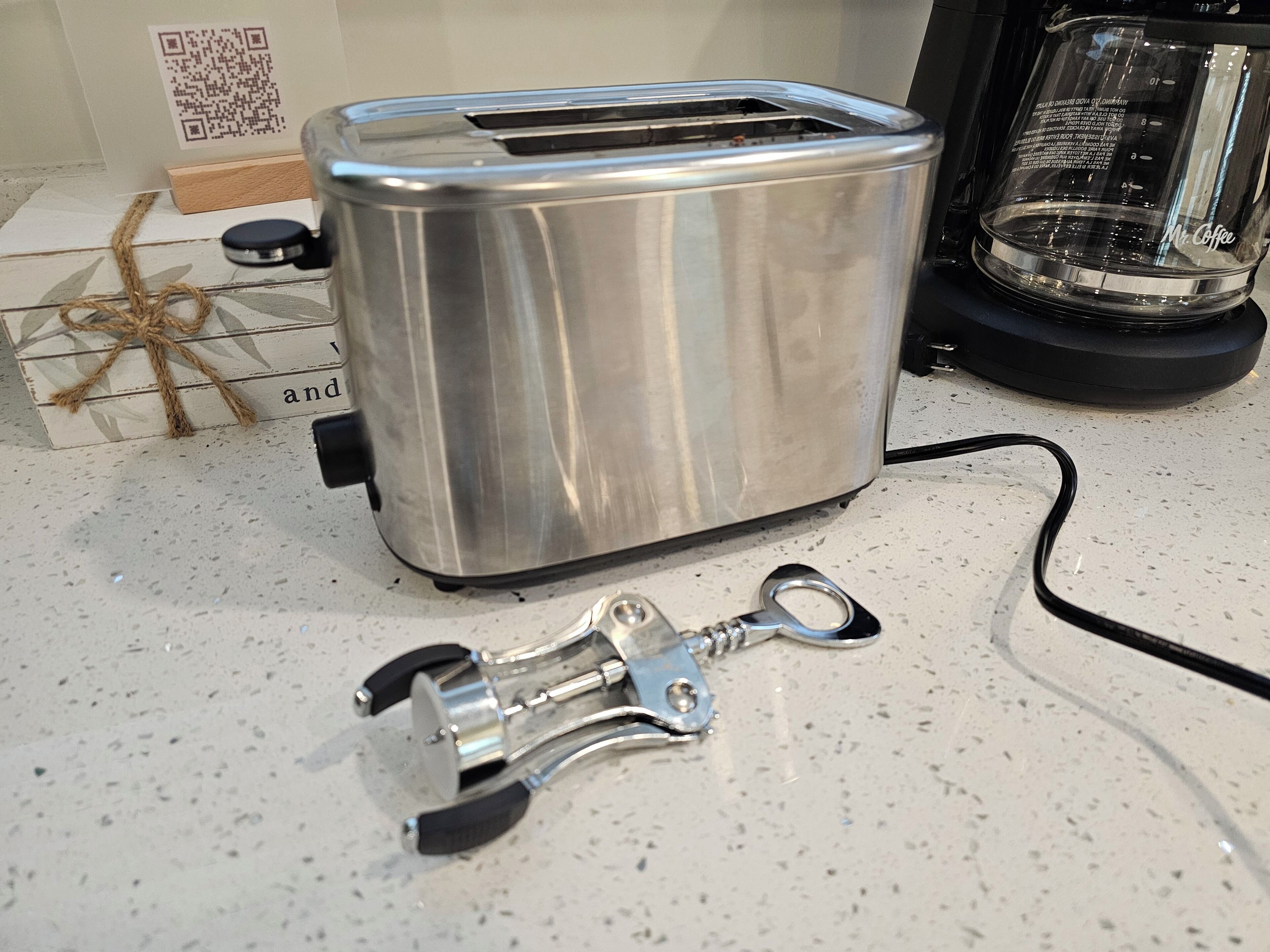 A stainless steel toaster on a kitchen counter with a QR code, a glass container, and a metal tool.