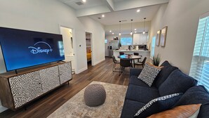 Un soggiorno moderno con una TV a schermo piatto che mostra il logo di Disney+, un divano blu scuro con cuscini decorati, un mobile TV in legno e un poggiapiedi rotondo.