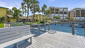 A wooden deck with a pool, palm trees, and a building with balconies.