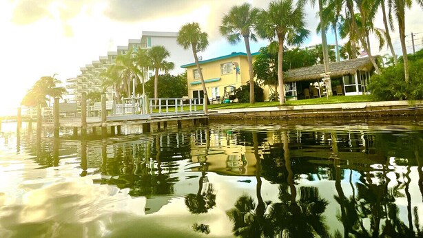 A waterfront property with a reflective pool, a thatched-roof structure, and a multi-story building with balconies.