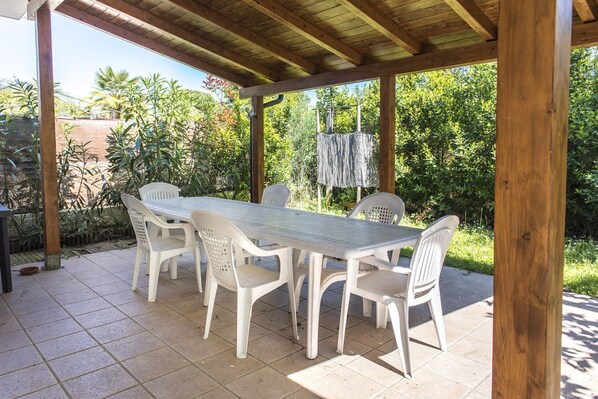 Eine überdachte Terrasse mit Tisch und Stühlen, umgeben von viel Grün.