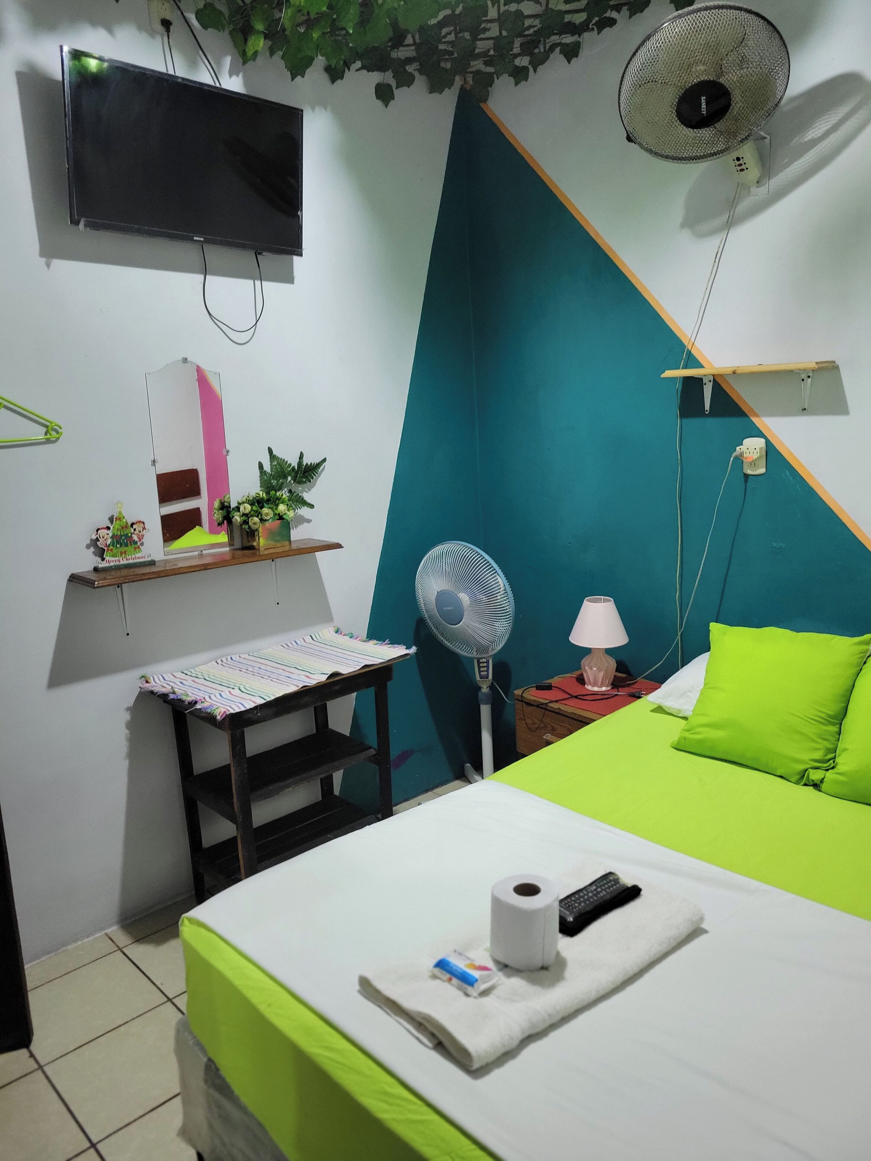A bedroom with a bed, a desk, a chair, a TV, a fan, and a bedside table with a lamp.