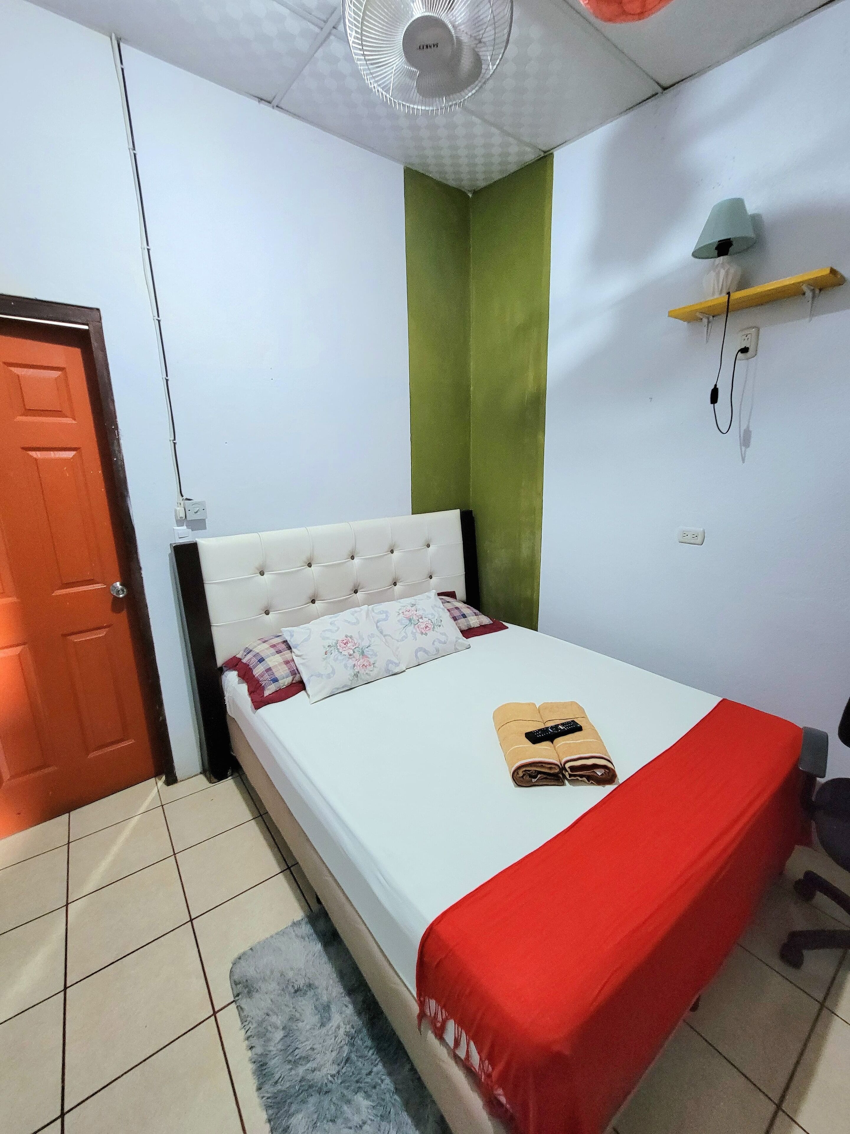 Une chambre à coucher bien rangée, avec un lit, une couverture rouge, une serviette pliée, une télécommande et un ventilateur au plafond.