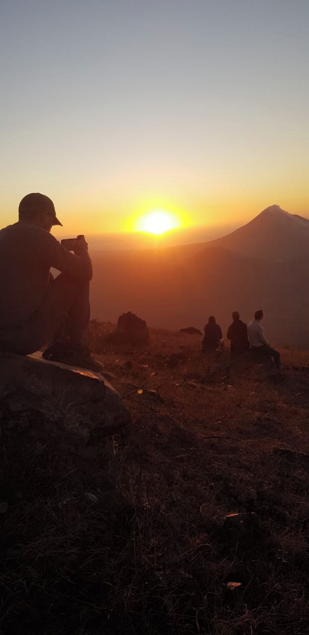 Eine Gruppe von Menschen sitzt auf einer Felsvorsprung bei Sonnenuntergang und blickt auf eine gebirgige Landschaft.