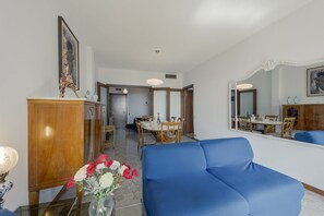 A living room with a blue sofa, a wooden cabinet, a dining area with a table and chairs, and a mirror on the wall. - GuestHost - Cozy apartment of about 110 square meters, able to accommodate up to 5 people, located on the 2nd floor of a building with lift and garage.The apartment has an enchanting view of the sea and the marina, surrounded by the greenery of Villa Orm (Sanremo)