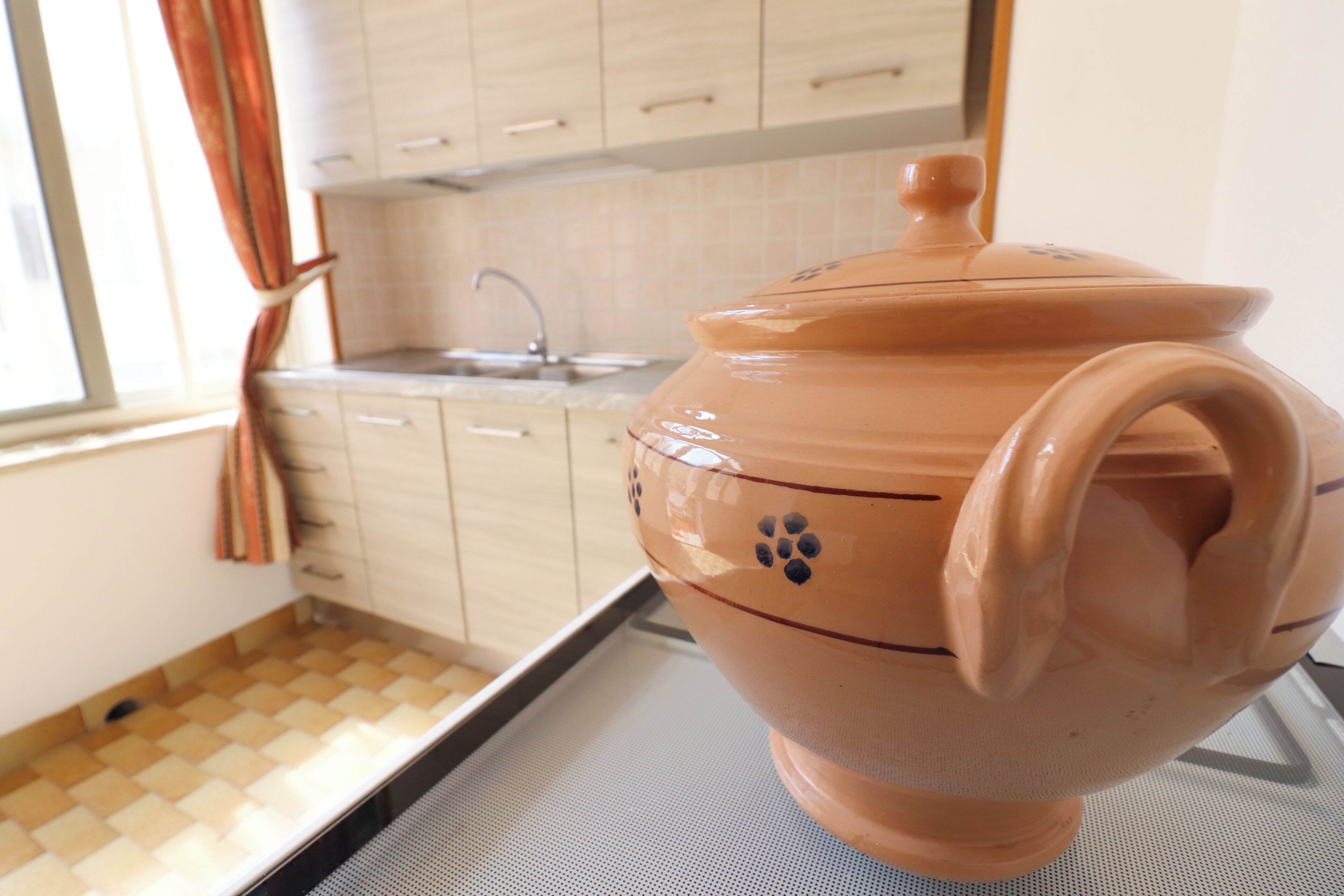 A kitchen with a large terracotta pot on a tray, a window with curtains, and a tiled backsplash.