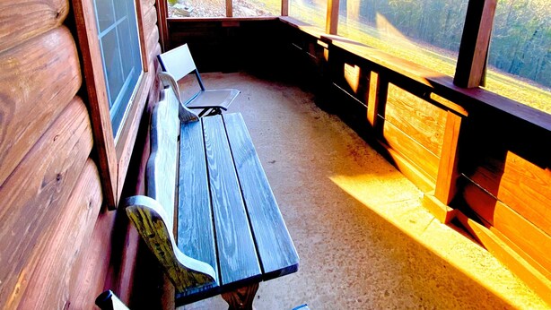 A covered porch with wooden benches and a table, large windows offering a view of a forested area.