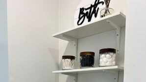 A bathroom with white walls, a toilet, and a shelf above the toilet holding jars and a black box.