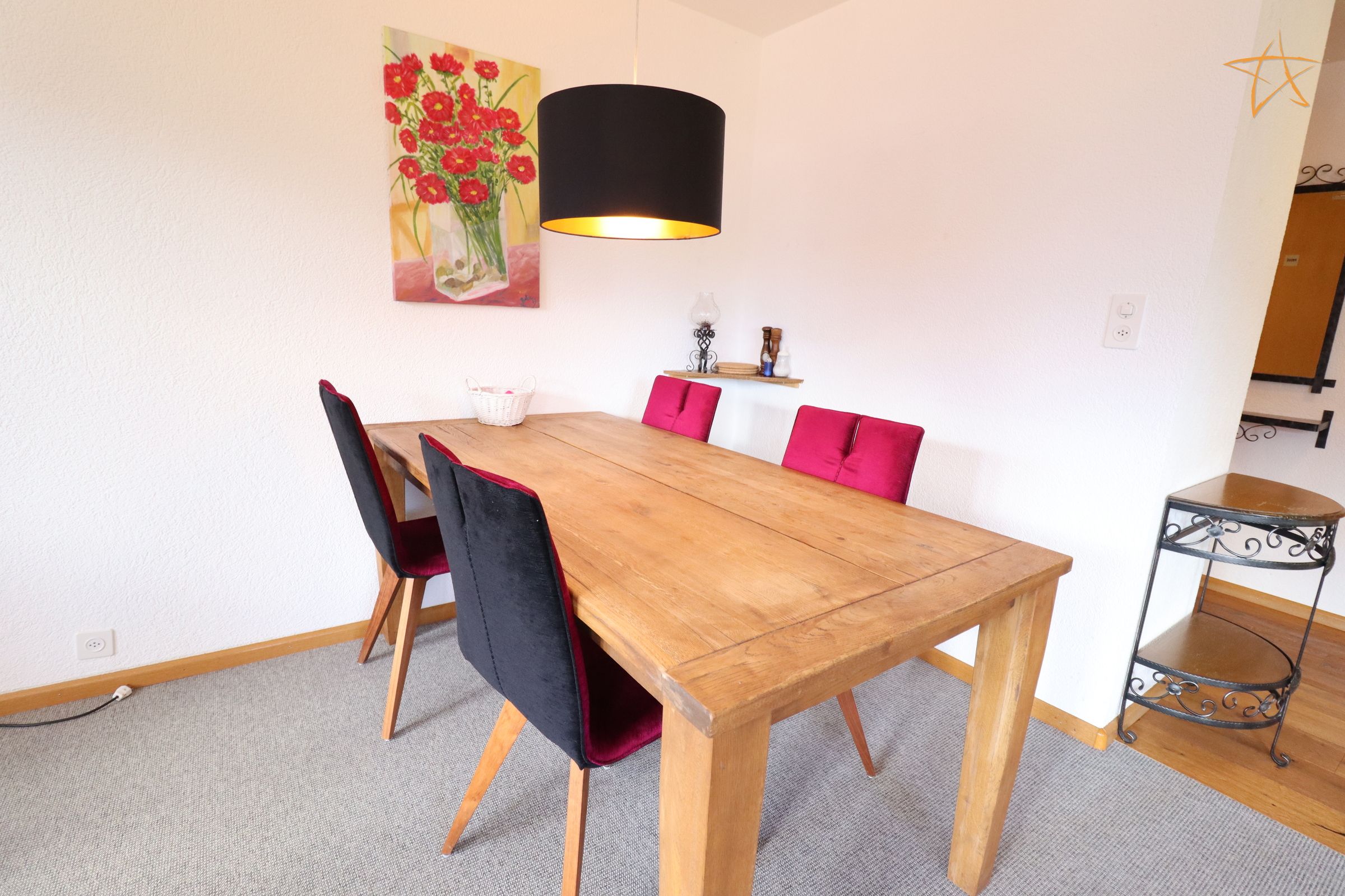 Un coin repas avec une table en bois, des chaises noires et rouges, une lampe noire et un tableau représentant des fleurs accroché au mur.