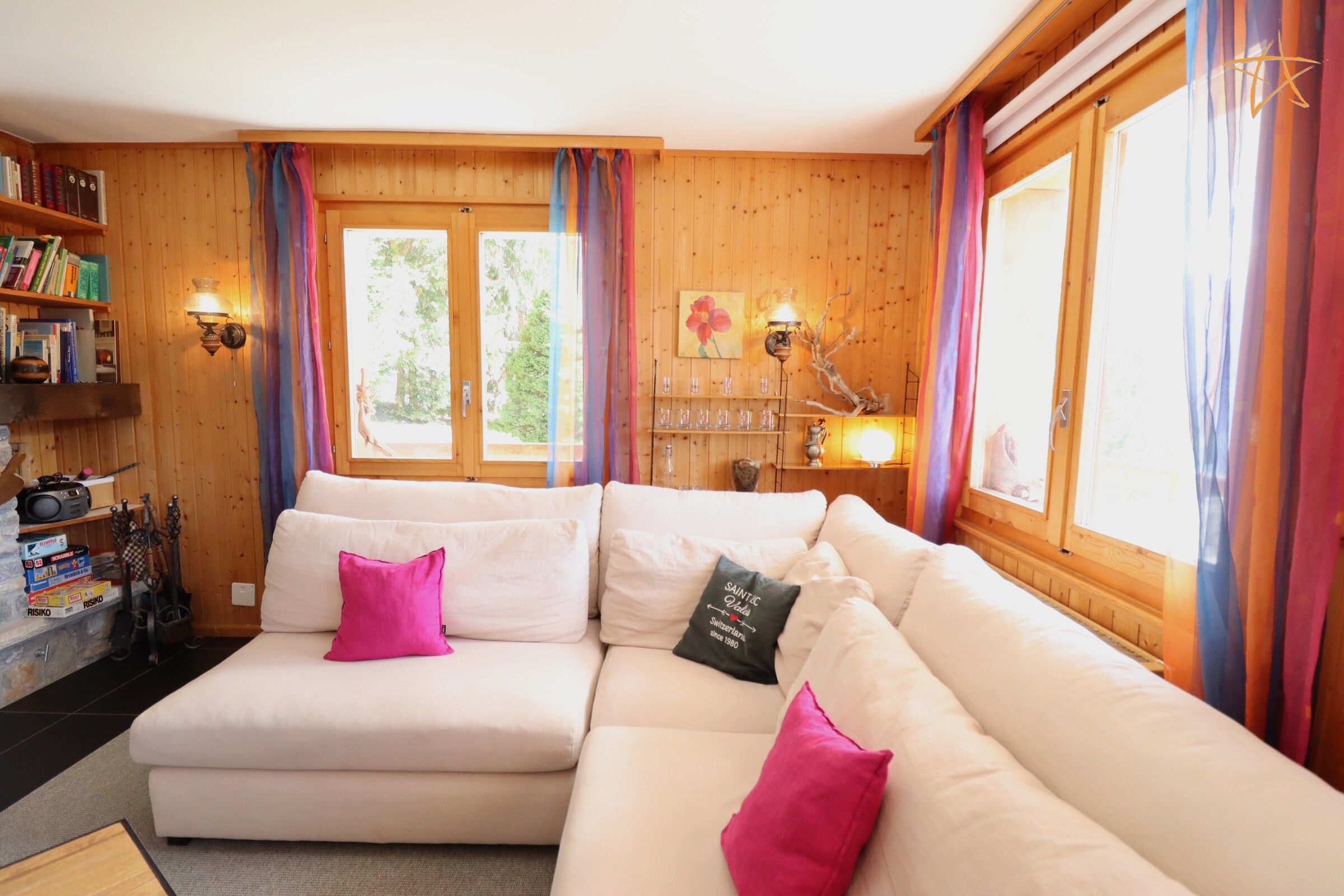 A cozy living room with a sectional sofa, a bookshelf, and a window with curtains.
