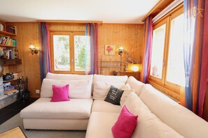 A cozy living room with a sectional sofa, a bookshelf, and a window with curtains.