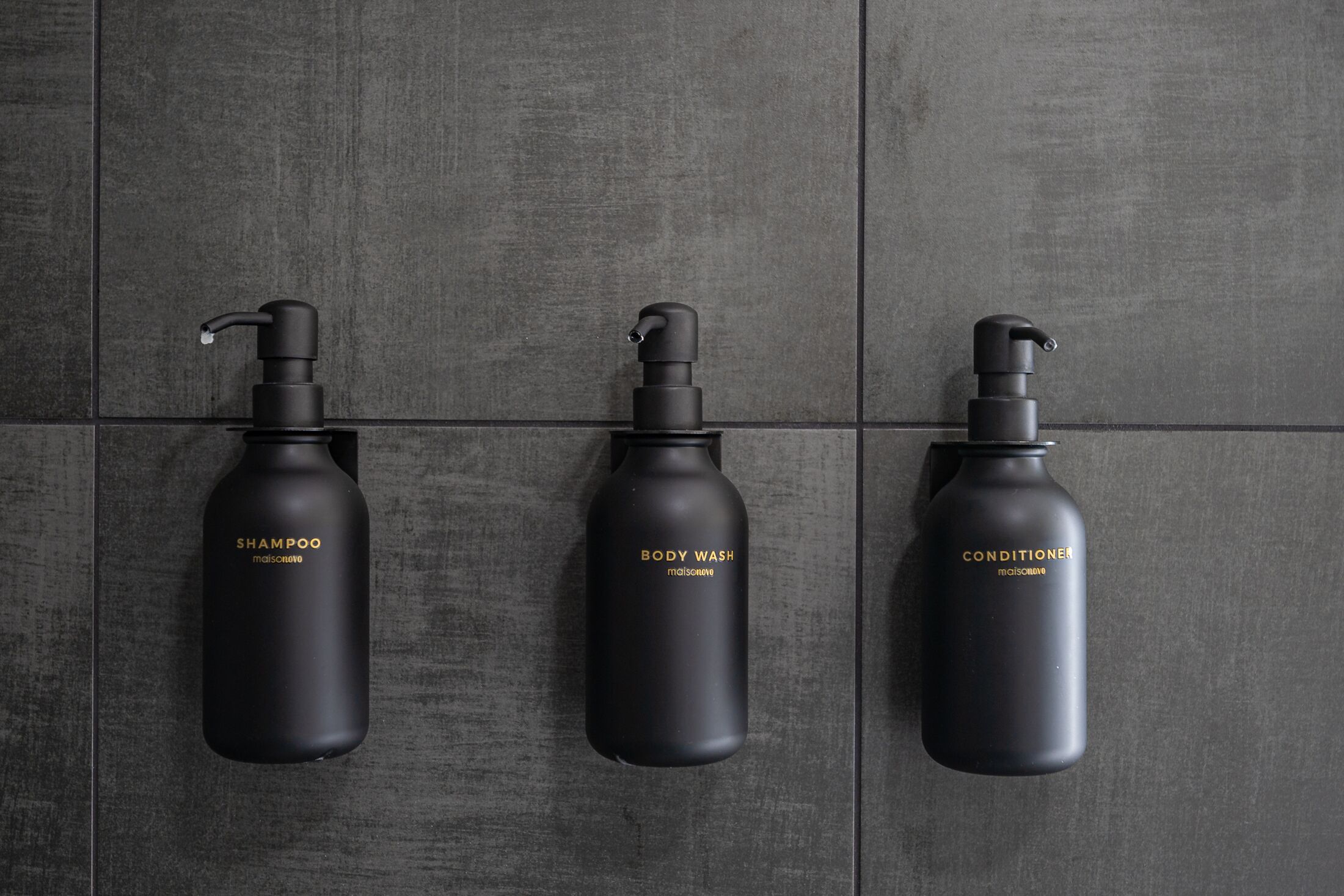 Three black bottles labeled with different toiletries (shampoo, body wash, conditioner) mounted on a tiled wall.