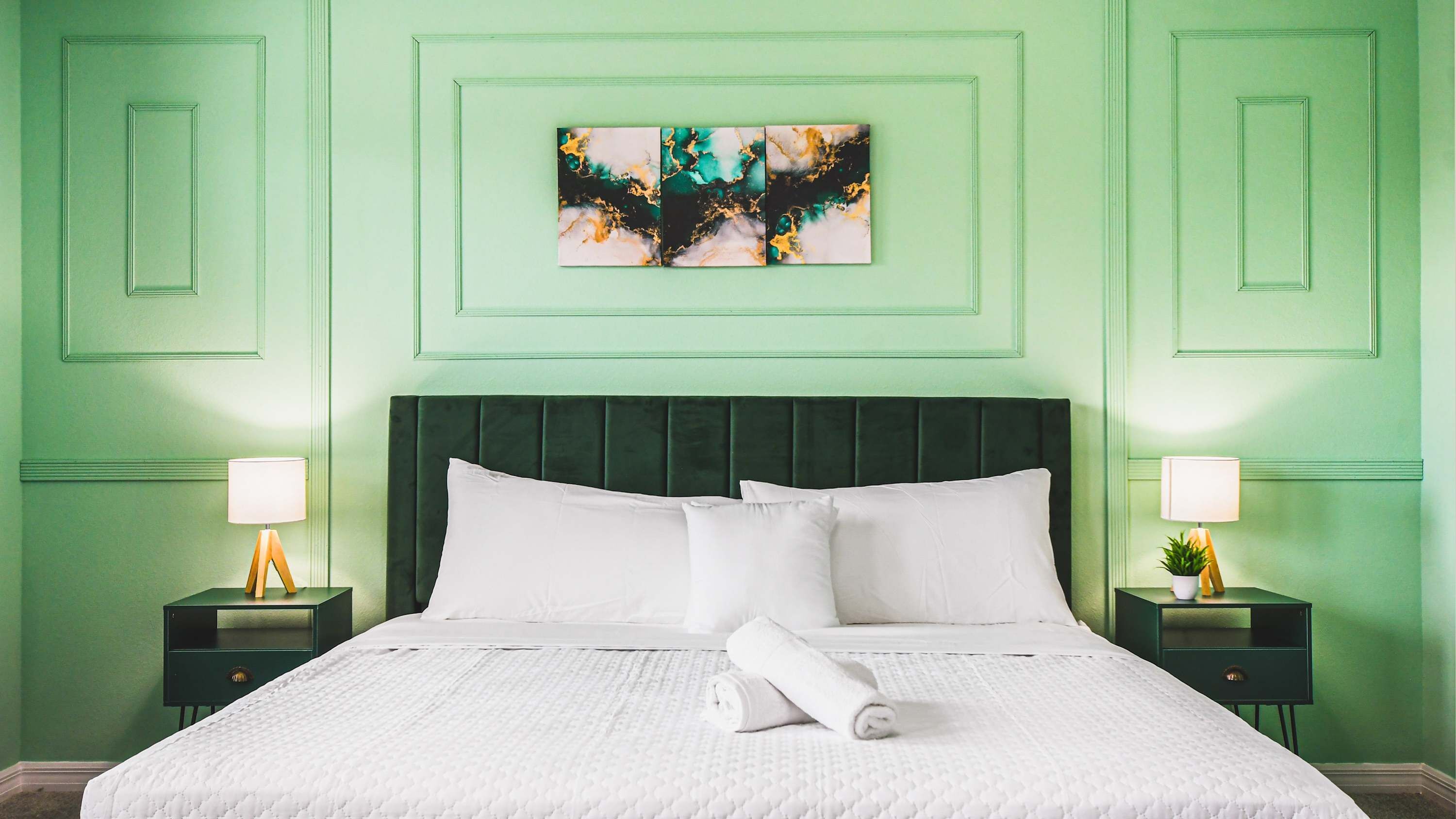 A neatly made bed with white linens and pillows, flanked by two bedside tables with lamps, in a room with green walls and a decorative abstract painting above the bed.