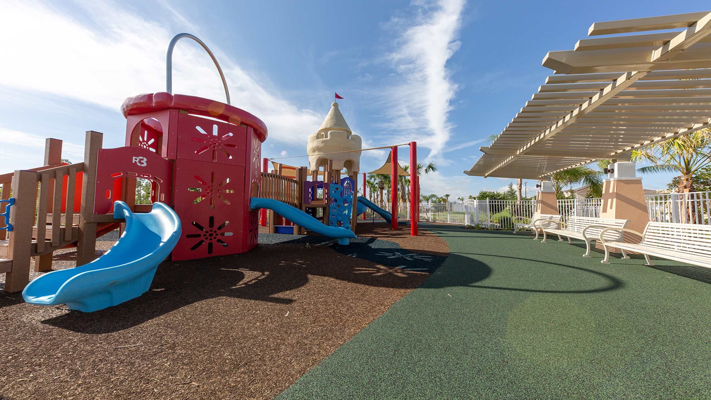 A playground with slides and climbing structures, surrounded by a fenced area with benches and a pergola.