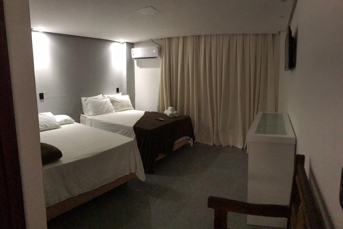 Hotellihuone, jossa on kaksi sänkyä, pieni pöytä, jossa on kaukosäädin, televisio ja verho.