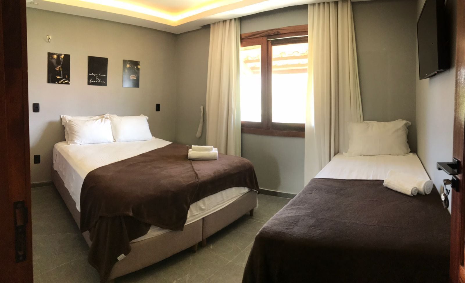 Ένα δωμάτιο ξενοδοχείου με δύο κρεβάτια, ένα μεγάλο παράθυρο με κουρτίνες και μια τηλεόραση επίπεδης οθόνης τοποθετημένη στον τοίχο.