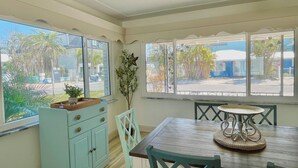 Un comedor con mesa y sillas de madera, un armario azul claro y ventanas grandes que dan vistas a palmeras y edificios.