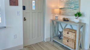 Un pasillo con puerta de madera, una consola con lámpara, un cuadro y cestos de mimbre.