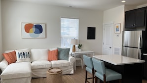 A modern kitchen with a white countertop and stainless steel appliances, a white sofa with patterned cushions, and a small dining area with two chairs. - ViBe District Retreat #2 (Virginia Beach)